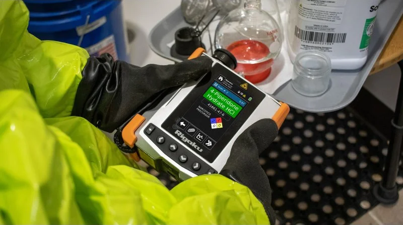 Responder in a yellow protective suit holding the CQL Max-ID with a 4-Piperidone Hydrate HCl result