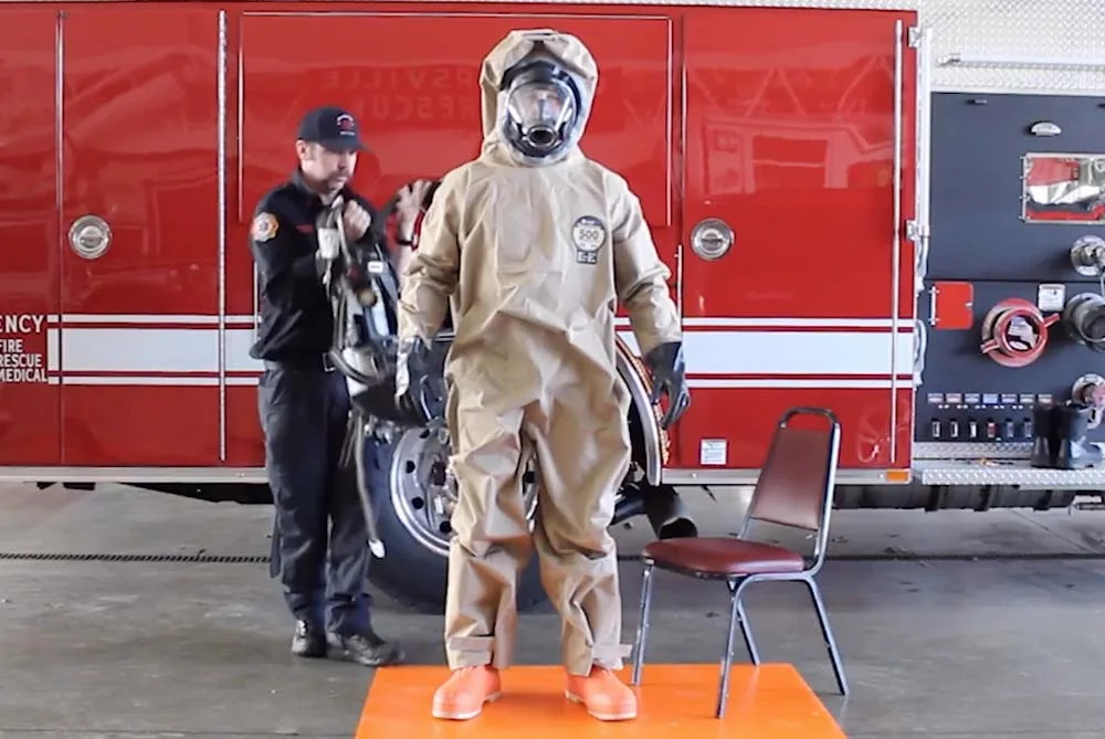 Responder standing in a DuraChem 500 suit on an orange mat while an assistant prepares the SCBA, in front of a fire engine