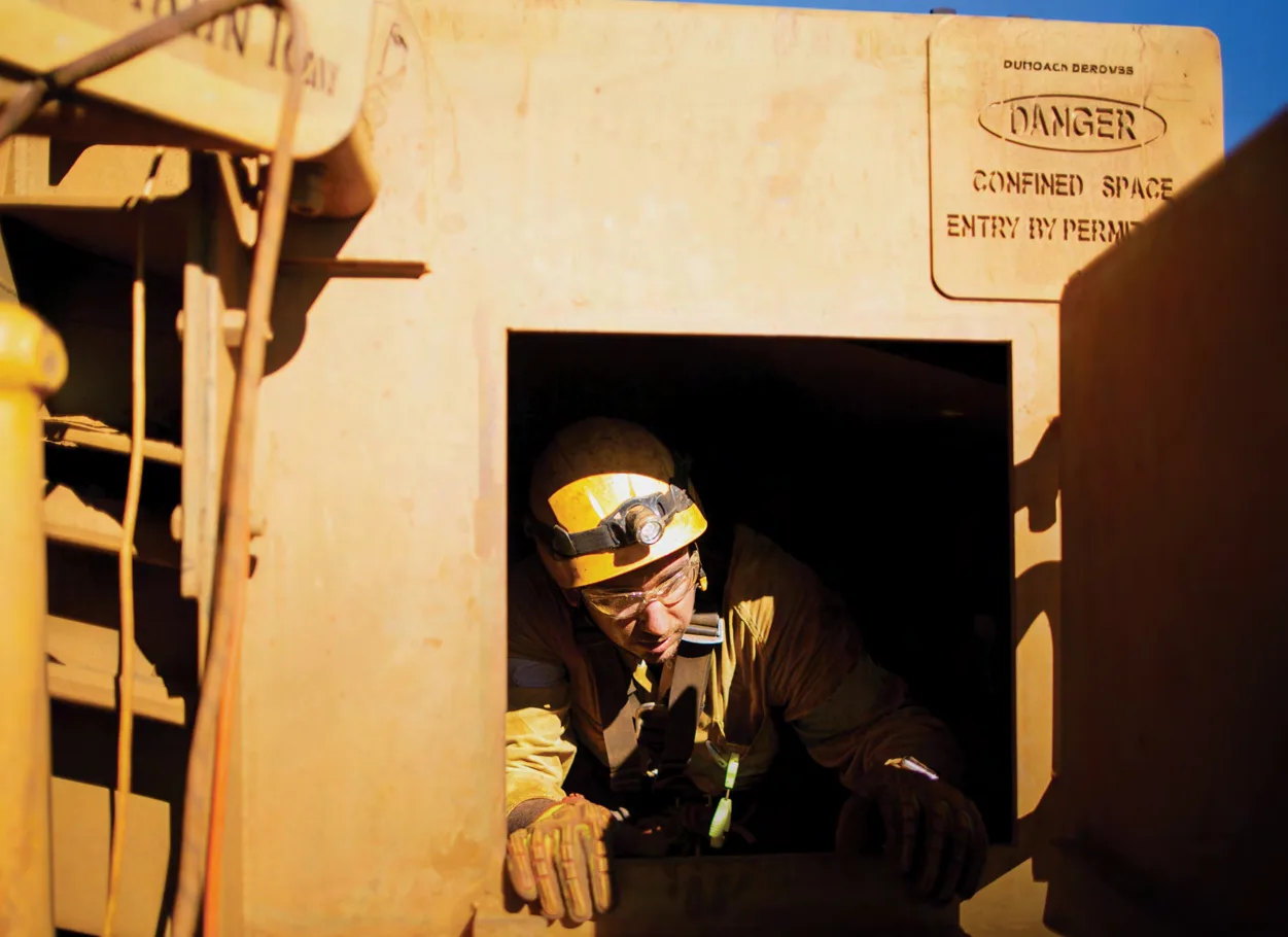 Worker in a hard hat and headlamp climbing out of a confined-space hatch beneath a stencilled sign reading Danger, Confined Space, Entry by Permit