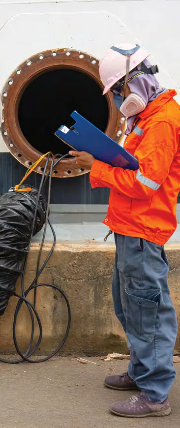 Worker in a respirator and orange coveralls reviewing a clipboard beside an open manway