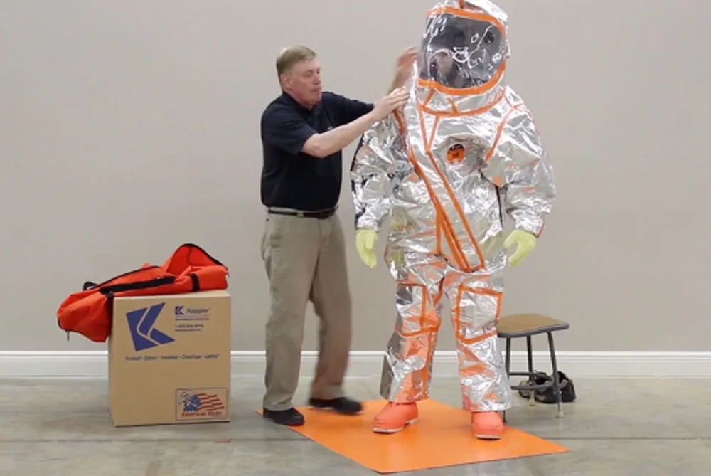 An assistant helping a responder don a silver Frontline 500 suit on an orange mat, with a carry bag and suit box beside them